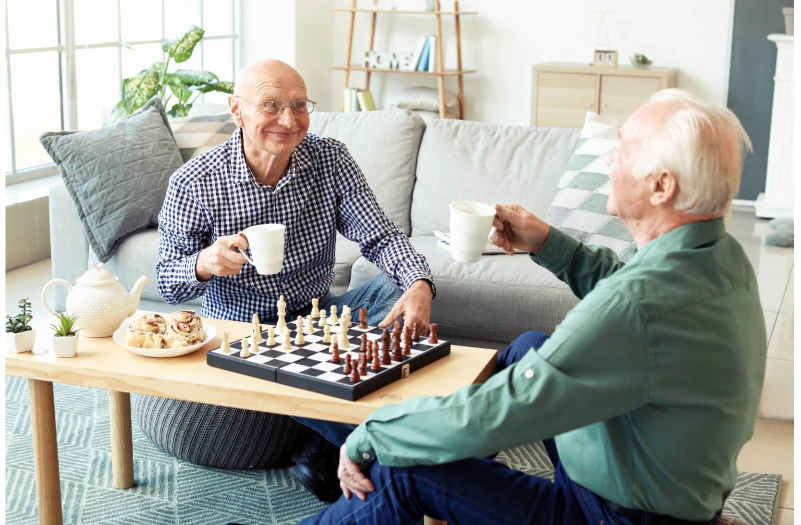 Two older adults sitting around a coffee table and playing chess while sharing a pot of tea and cinnamon rolls