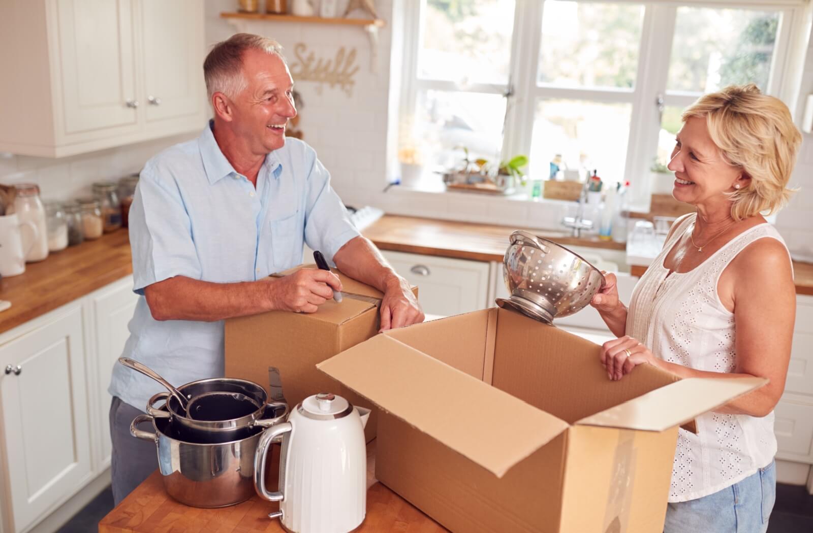 An older adult couple putting kitchen items ina cardboard box.