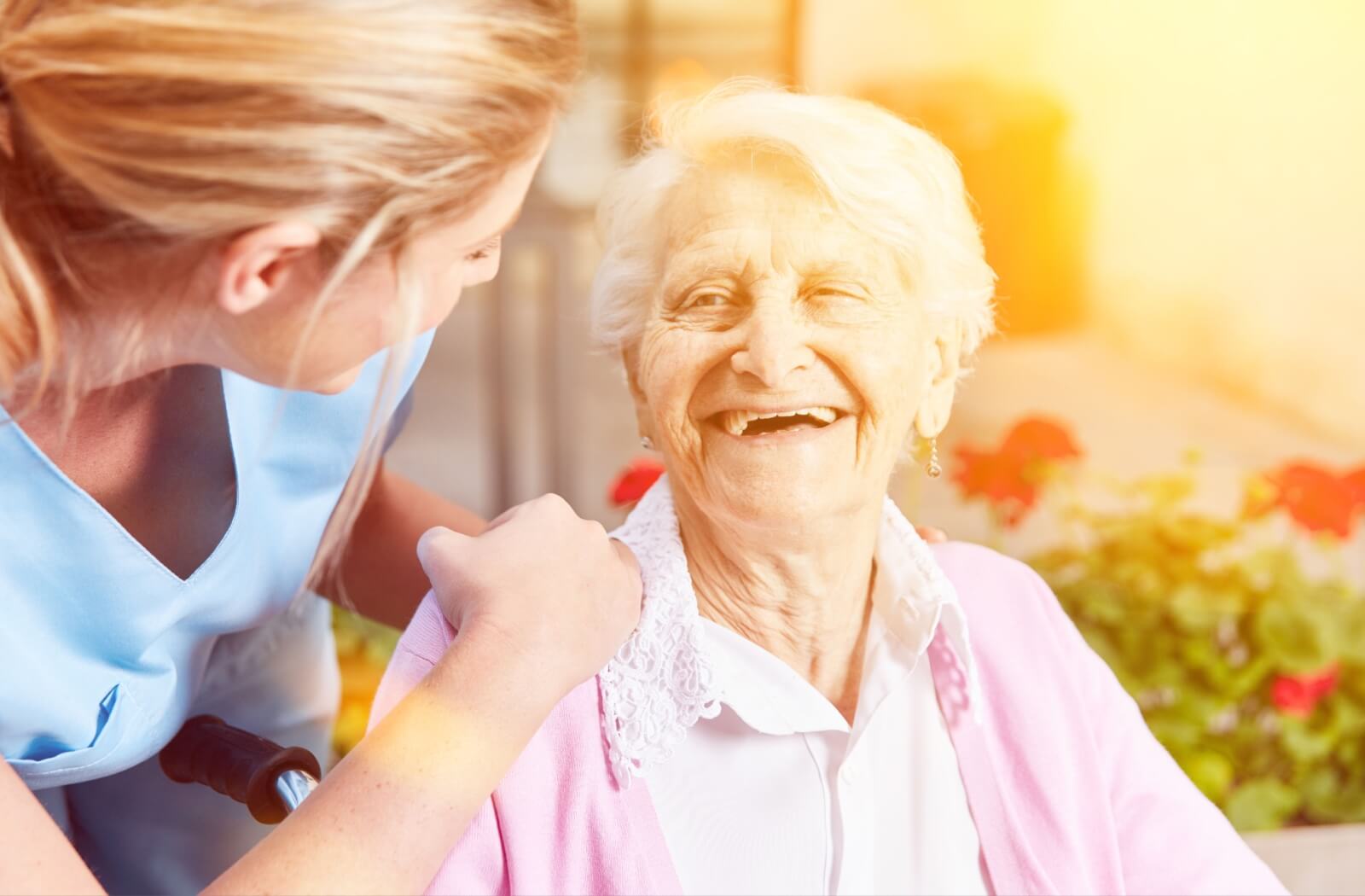 An older adult woman in a senior living facility sitting on a chair smiling and having a conversation with a nurse.