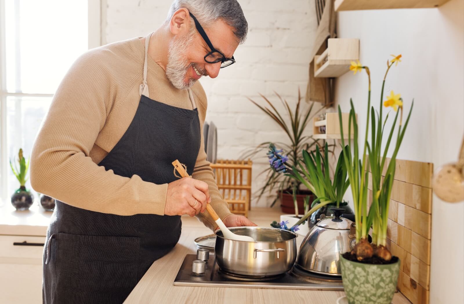 An older adult man in a black apron preparing a meal in a sunlit kitchen.