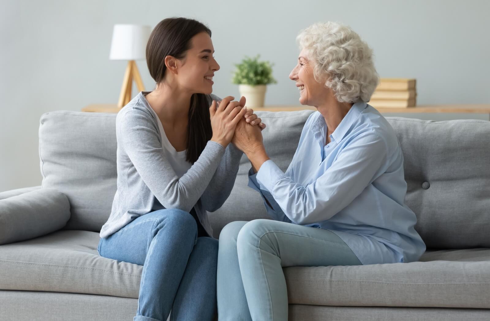 A loving daughter and her older adult mother sitting on a couch holding hands and smiling.
