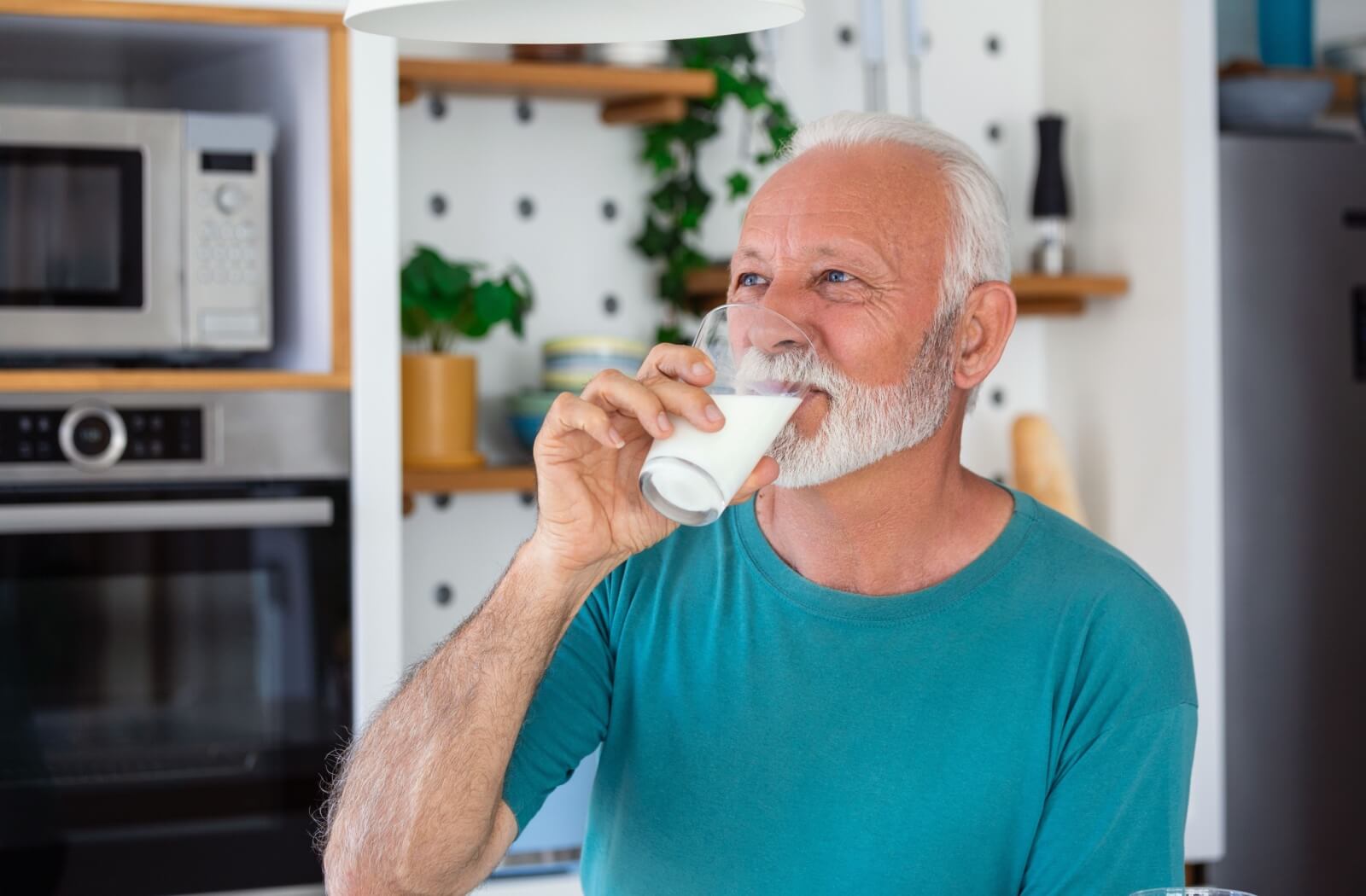An older adult man drinking a glass of milk in his kitchen.