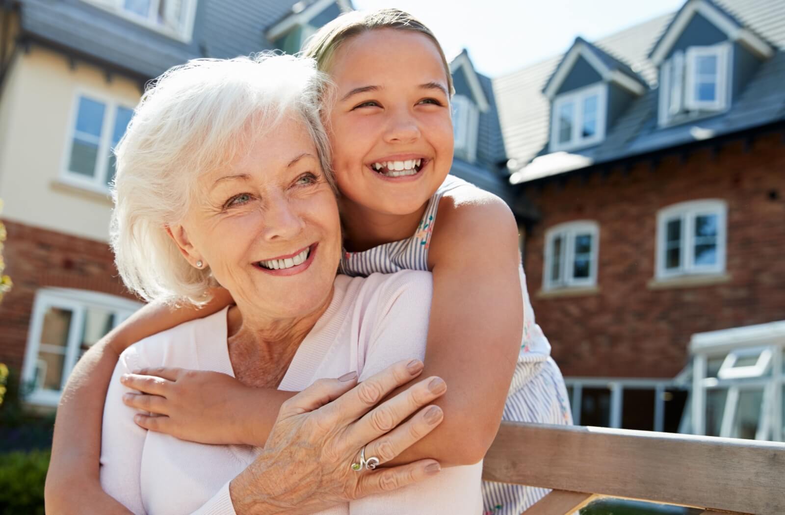 An older adult woman and her granddaughter are hugging and smiling on an outdoor bench.