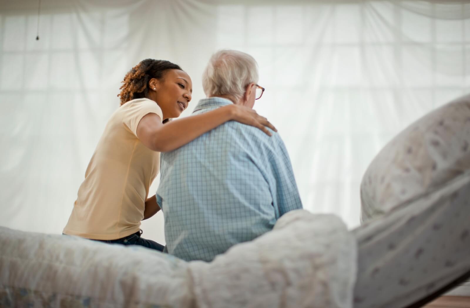 A care assistant comforting a senior man in pajamas, who is seated on his bed and expressing a desire to leave the assisted living community.