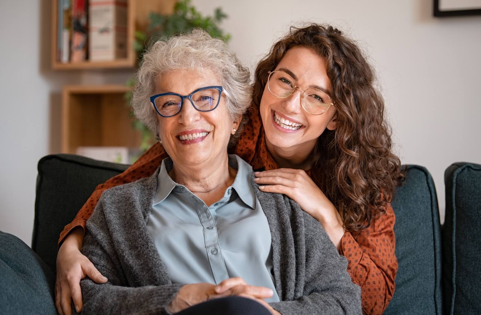 A young woman warmly hugging her elderly mother from behind.