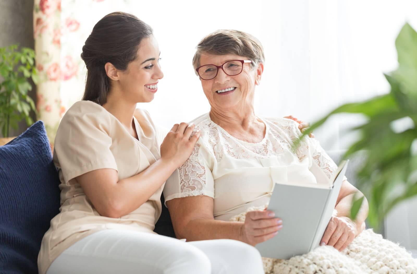 A caregiver sitting alongside a joyful elderly woman in glasses, who is engaged in reading a book about Alzheimer's.