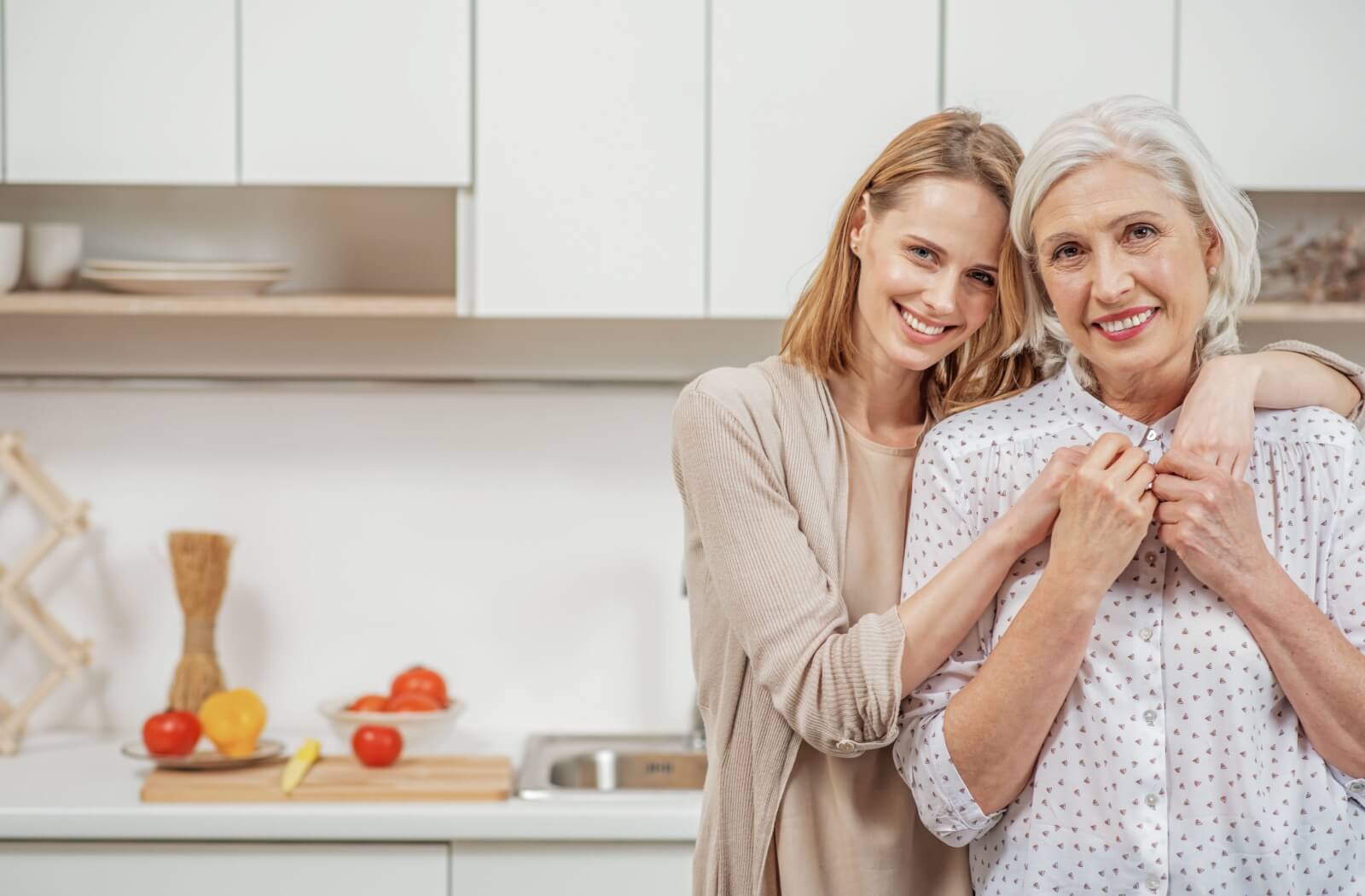 An older adult woman and her daughter smile and hug in a kitchen.