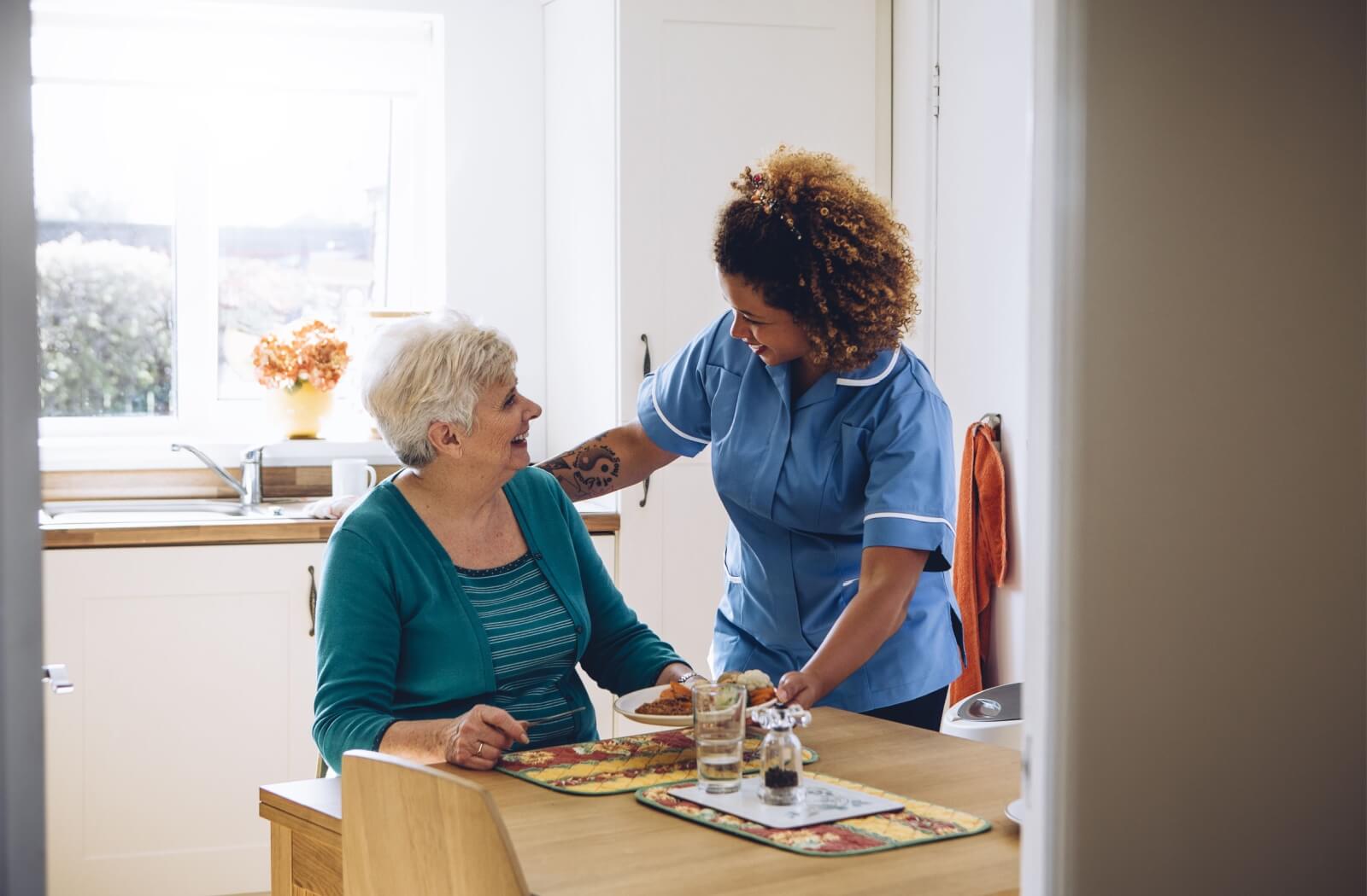 A smiling assisted living community staff member delivers a delicious meal to a resident at her kitchen table.
