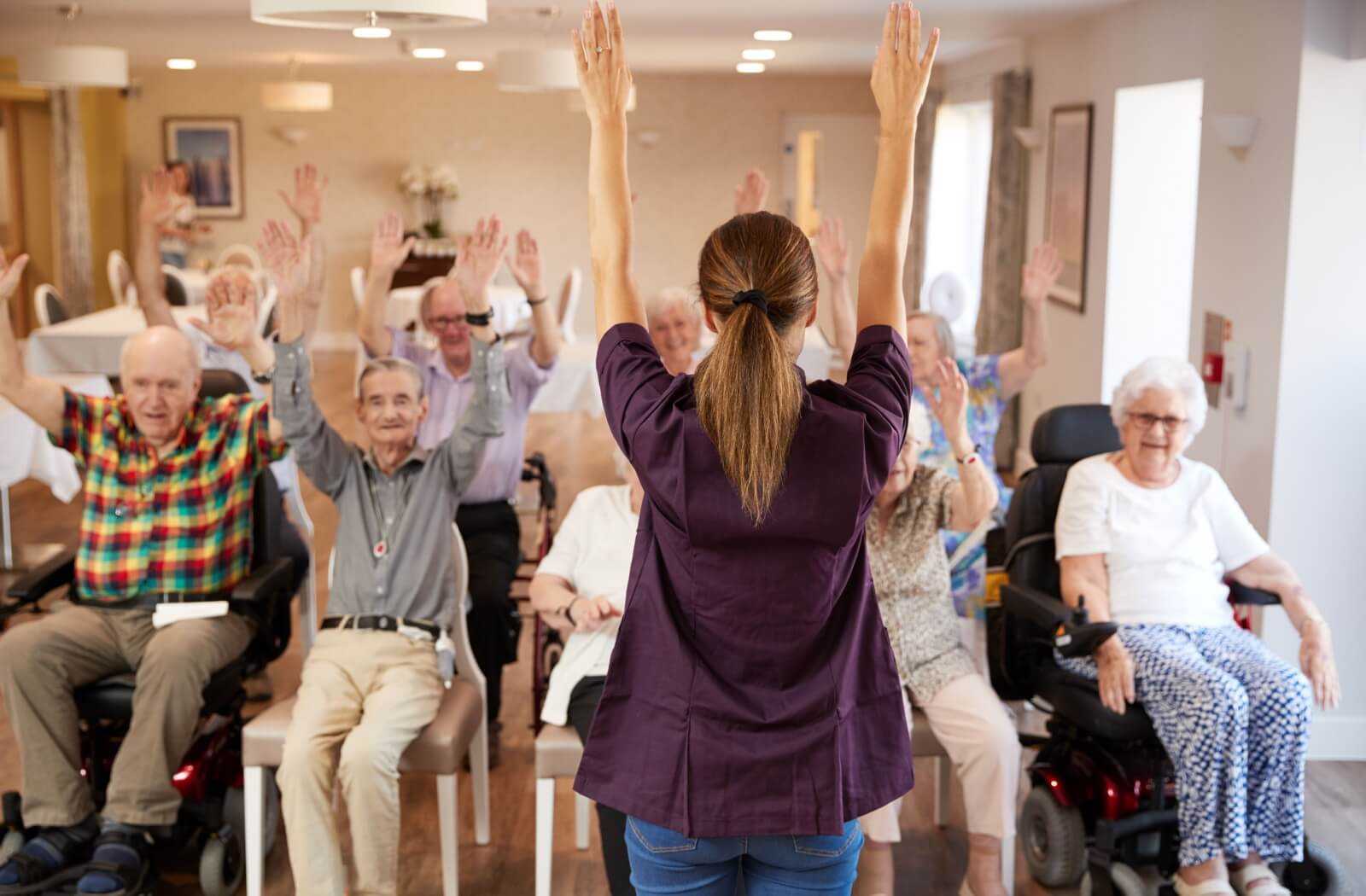 A group of older adults in an assisted living community performing stretching exercises led by a staff member.