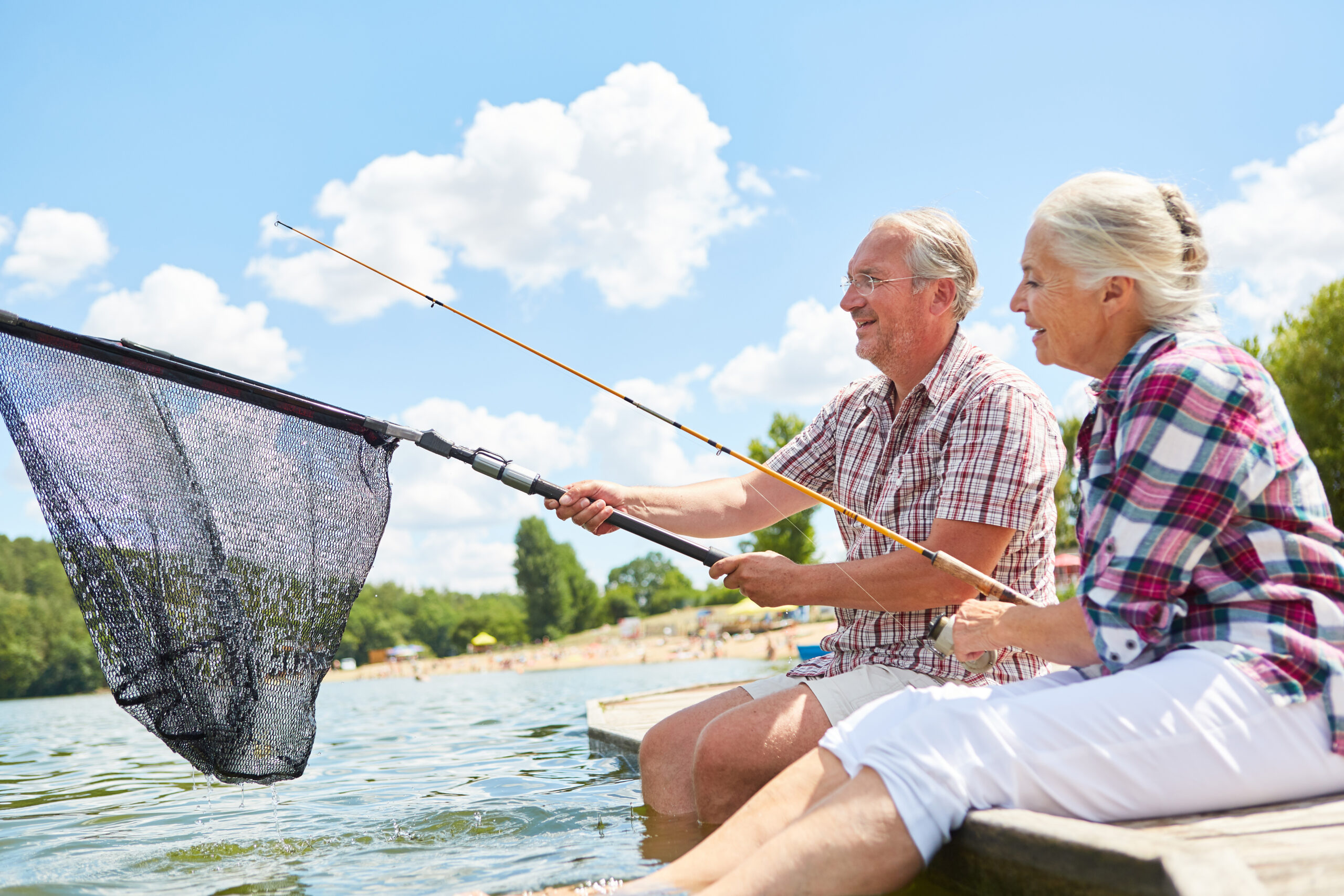 An older adult couple fishing together on a dock on a sunny day.