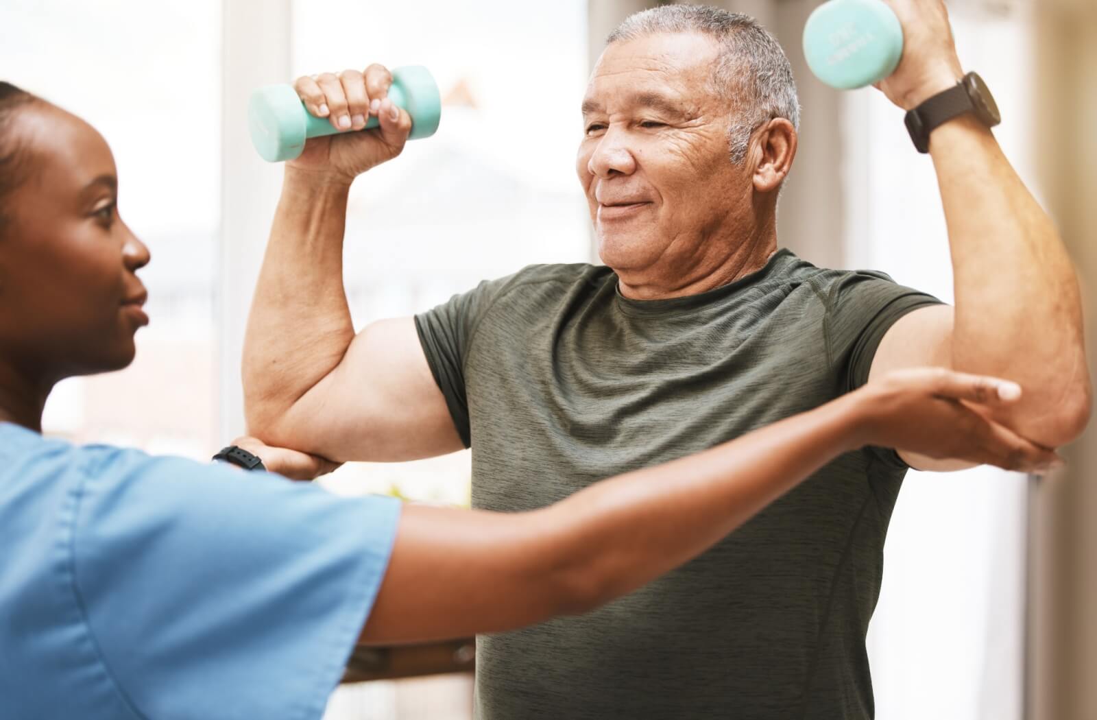 A young physical therapist helps an older adult man lift weights as part of his physical therapy.