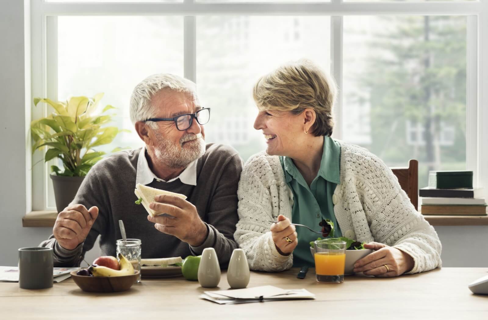 An older adult couple sit together in the kitchen, smiling and eating whole foods.