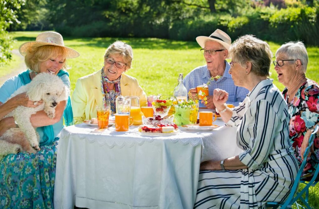 An older adult woman playing with a dog at a park with other older adults and sitting at a table enjoying iced tea.