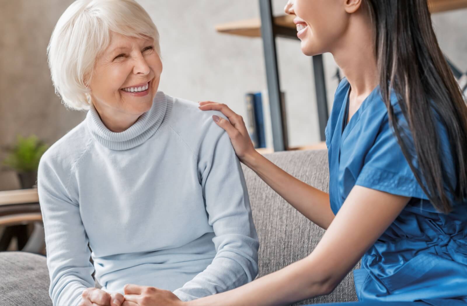 A caregiver and memory care resident smile at each other while sitting on the couch.