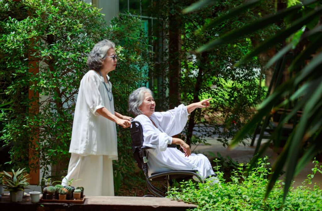 Two mature women wander through a memory care garden, one in a wheelchair.