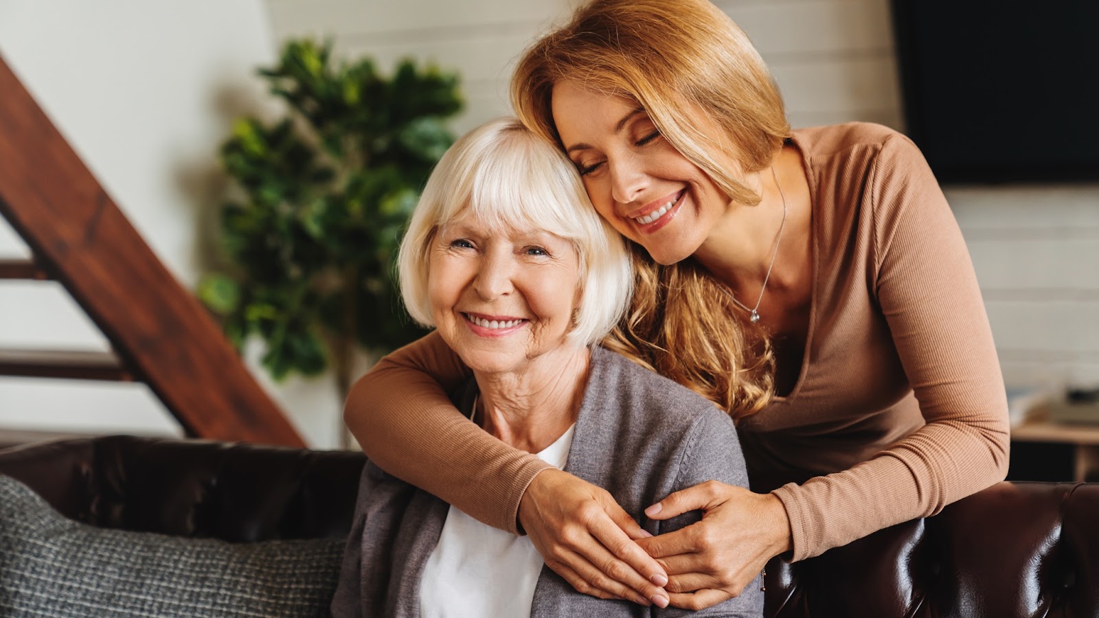 A happy mature daughter embraces her older adult mother from behind.