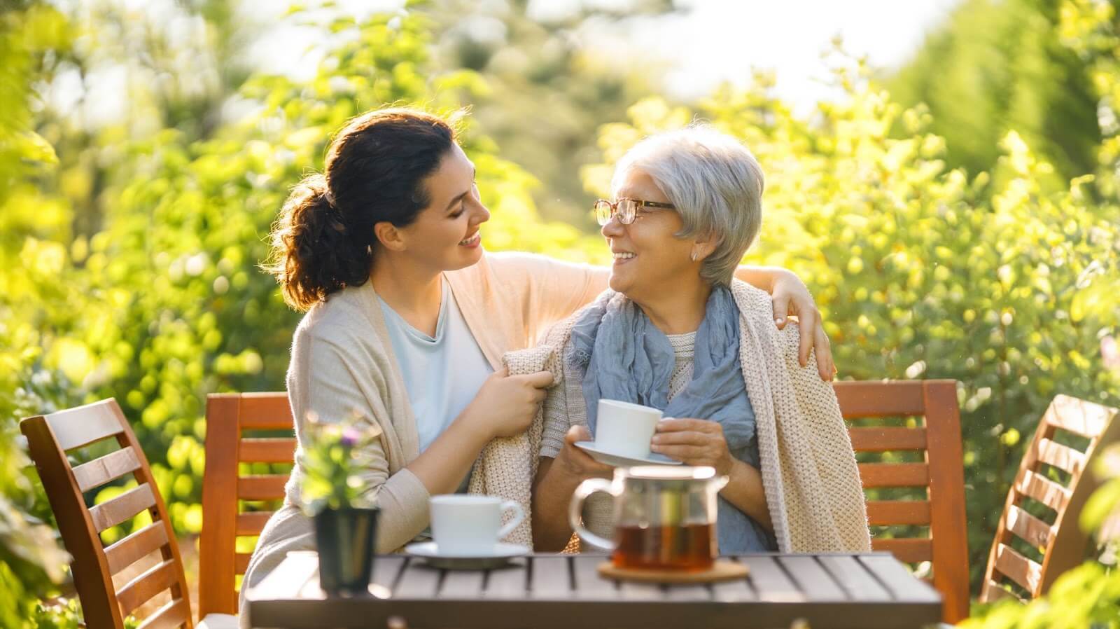 An adult daughter puts her arm around her mother while sitting at an outdoor table drinking tea.