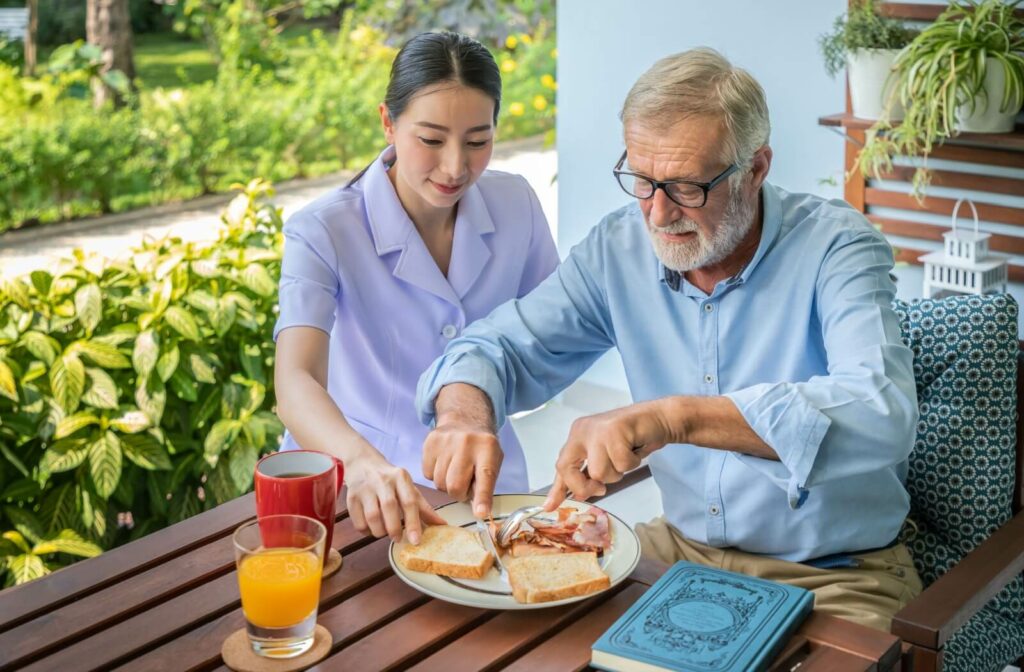 A senior living community caregiver assists an older adult with breakfast on the patio on a sunny day.