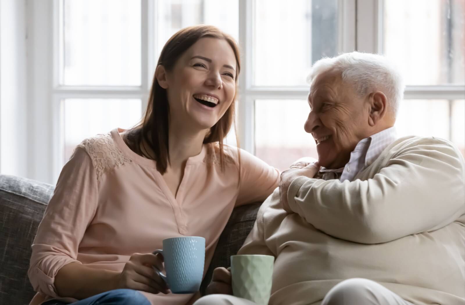 An adult child laughs with their parent over coffee in a cozy living room.