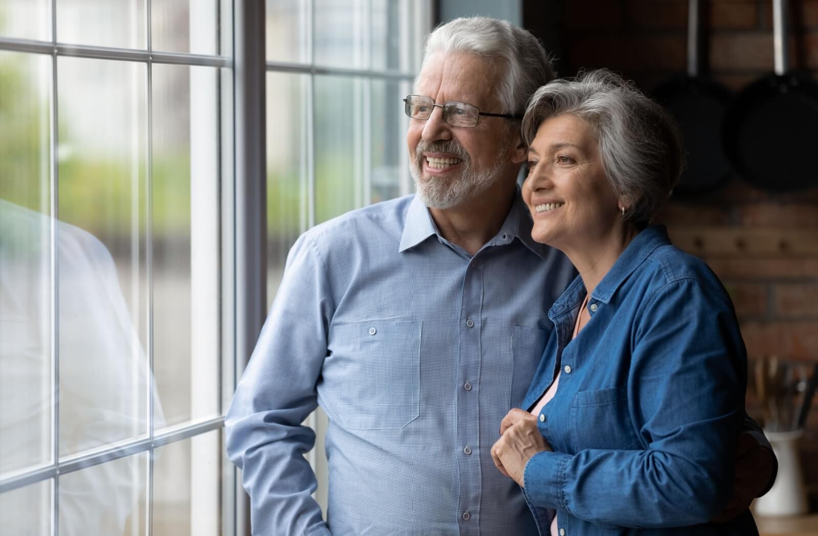 A mature couple embraces while looking out of the window in their independent living home.