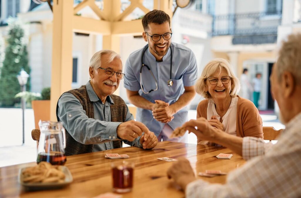 A group of older adults plays cards at a table with a caregiver nearby, laughing and enjoying the moment.