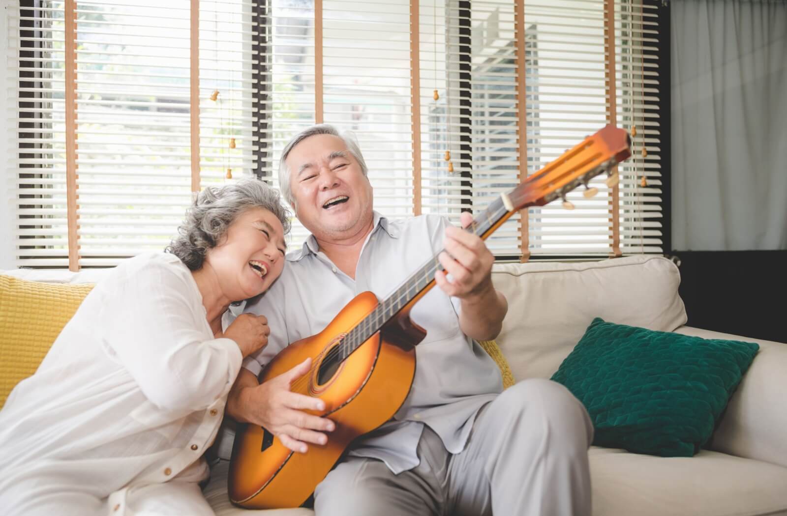 Two older adults laugh and sing together while one plays a guitar.
