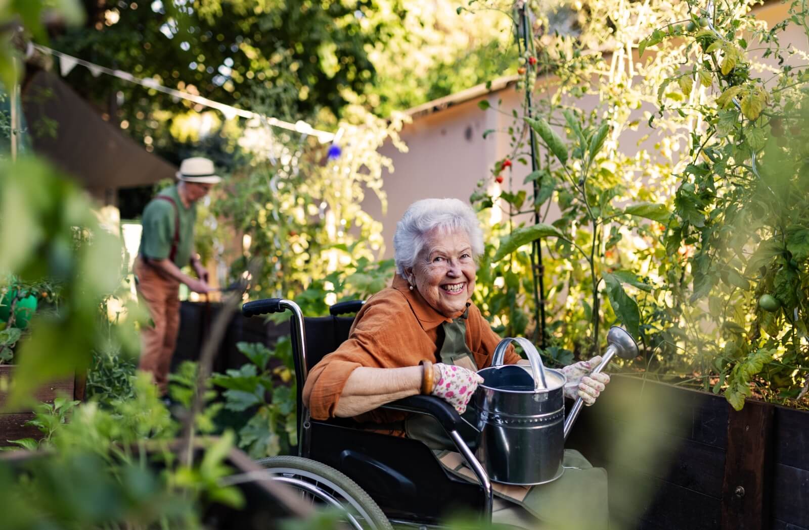 An older woman in a wheelchair holds a watering can and smiles in a garden. An older man can be seen in the background.