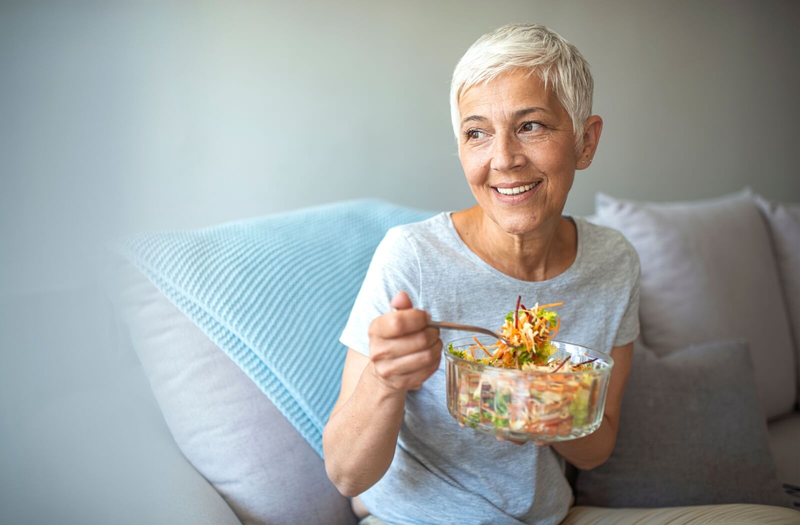 A smiling older adult sits on a couch and enjoys a bowl of salad.