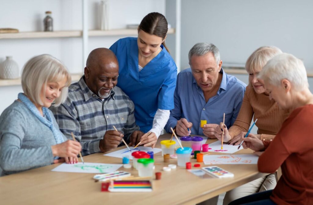 A group of older adults participate in painting together, while an assisted living staff member assists them.
