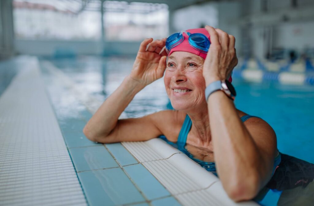 A happy older adult wearing a swimming cap adjusts their goggles at the side of the pool.