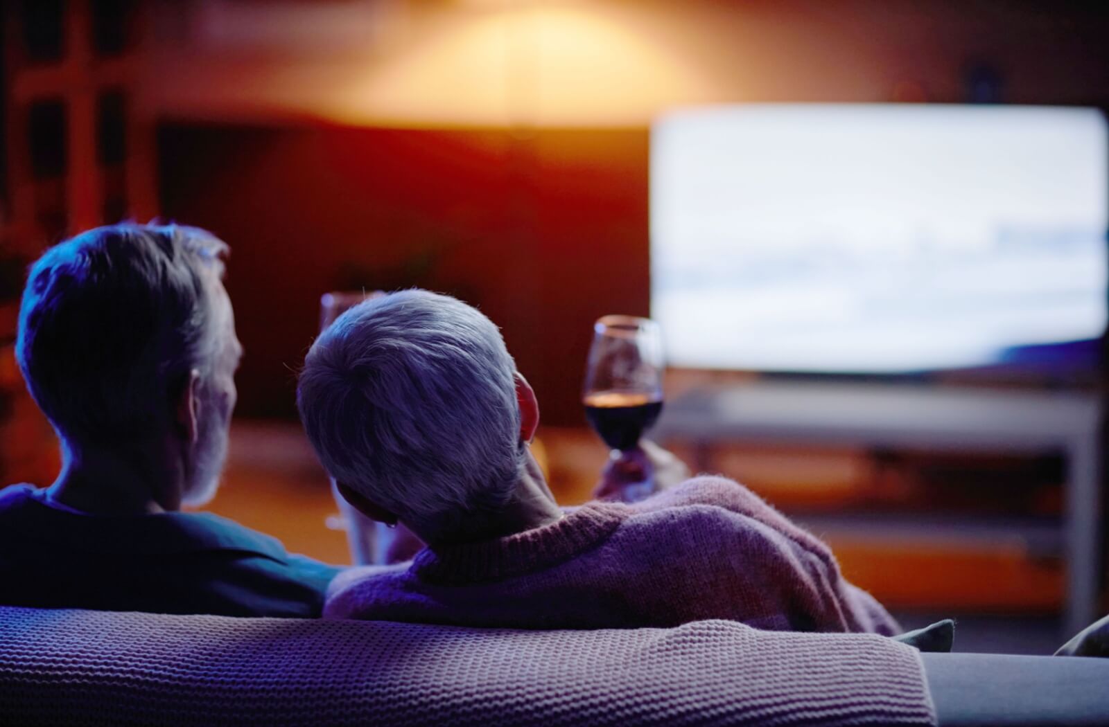 Two older adults sitting comfortably, enjoying a movie together with glasses of wine.