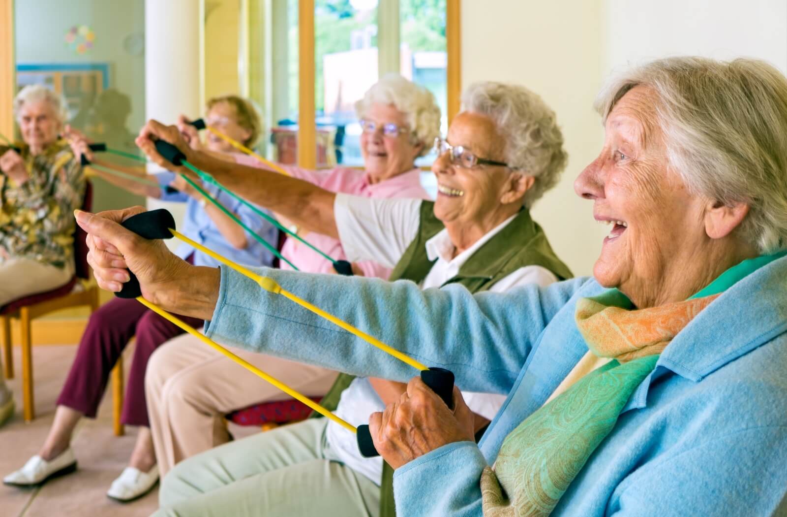 A lively group of older adults shares smiles and laughter as they move through a band exercise together.