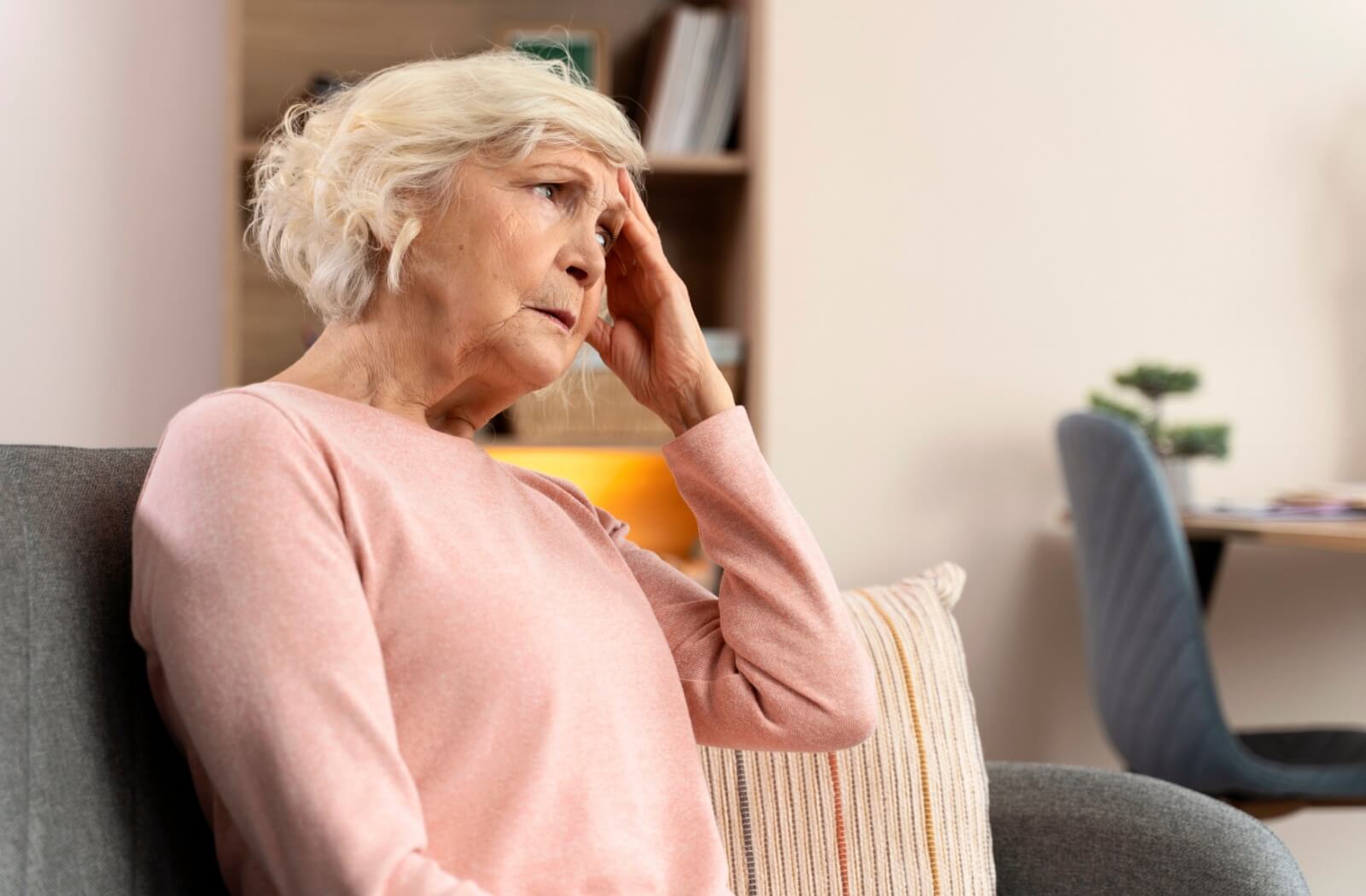 An older adult sitting on a couch, touching their forehead with a concerned expression, struggling with memory or cognition.