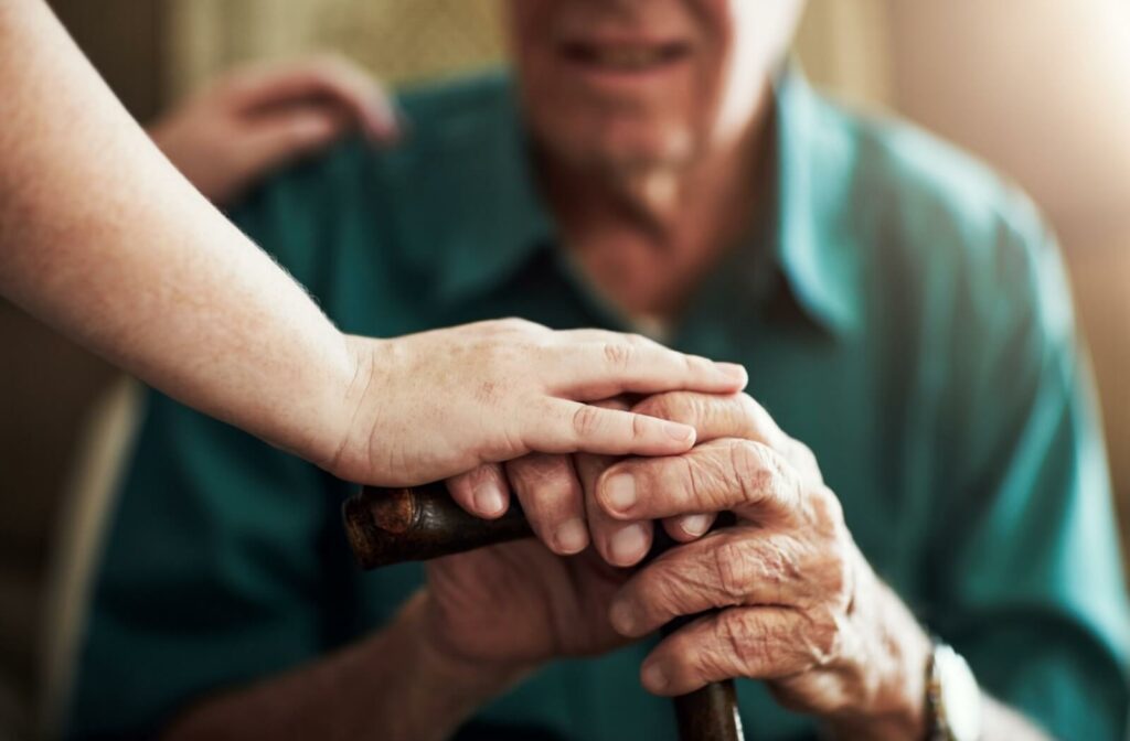A close-up of two hands, one gently placed over the other, offering comfort and support to an older adult holding a cane.