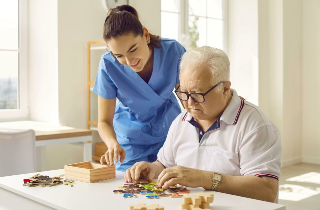 A caregiver in blue scrubs smiles while assisting an older adult with a puzzle in a bright, welcoming space.