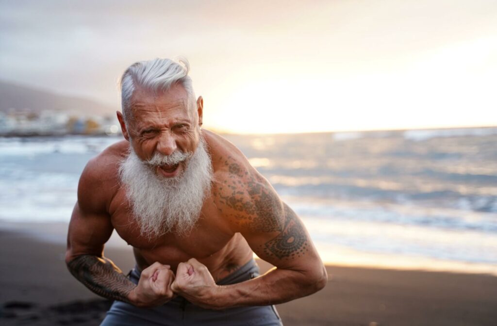 An older adult flexes his tattooed arms on the beach.