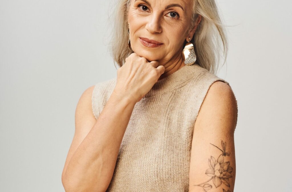 A graceful, mature woman strikes a pose. Her right hand rests under her chin, showcasing a striking tattoo on her left arm.