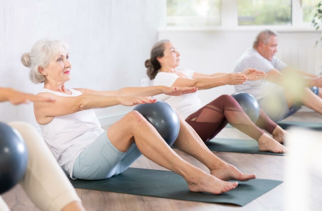 A group of older adults in a Pilates class perform seated exercises on mats with stability balls, focusing on core strength.