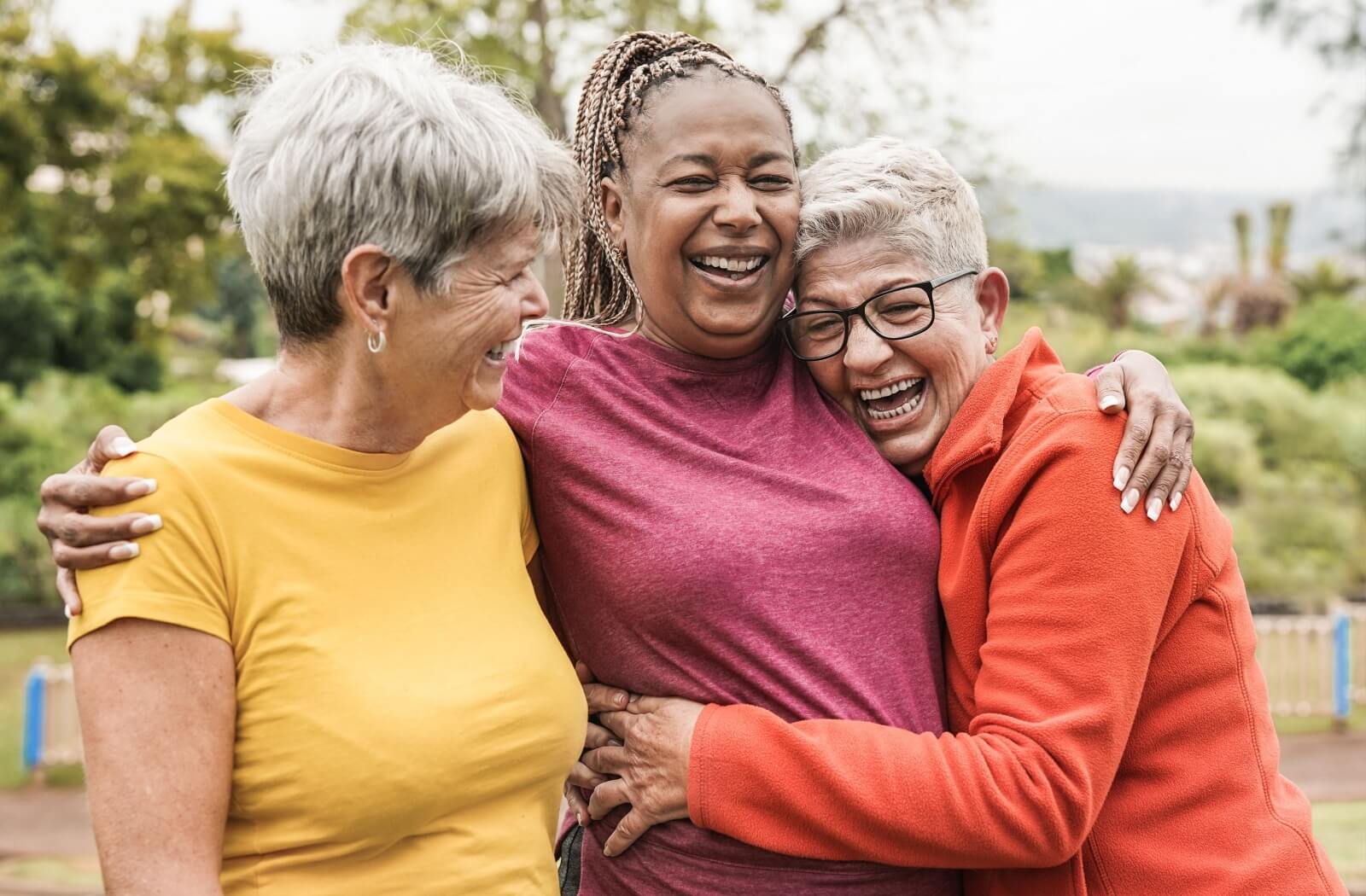 Three older adult friends laugh and embrace each other warmly in a park.