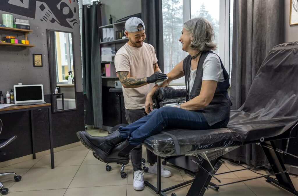 An older adult sits in a tattoo studio, smiling and chatting with a tattoo artist who is preparing to work on their arm.