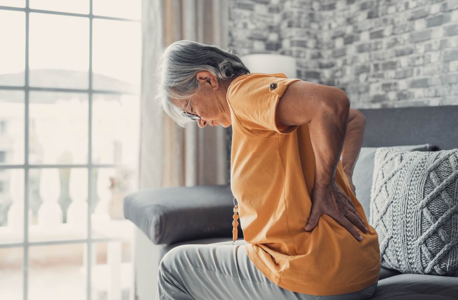 An older adult sitting on a couch, holding their lower back in discomfort, experiencing common age-related aches and pains.