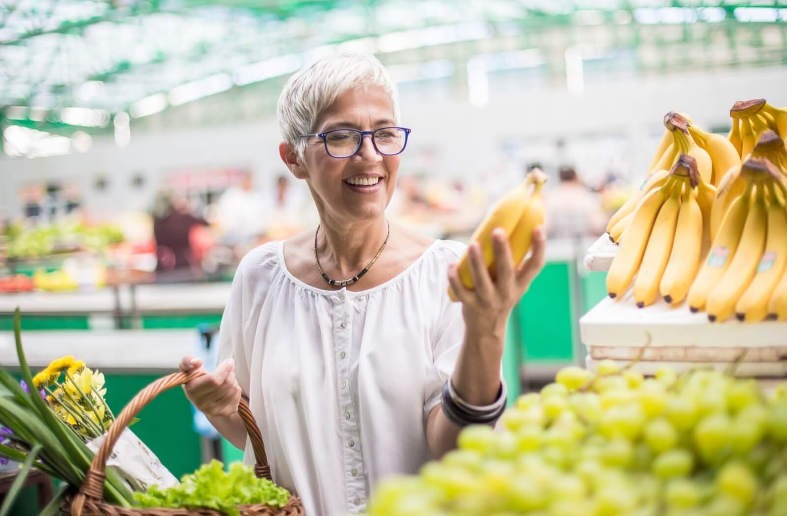 A mature woman at the grocery store picks up a bunch of bananas and inspects them approvingly.