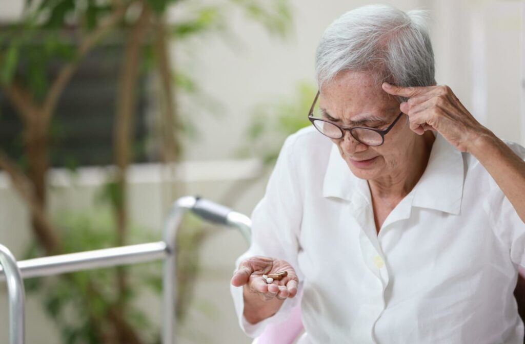 An older adult looking at a handful of pills with a concerned expression, touching their forehead.