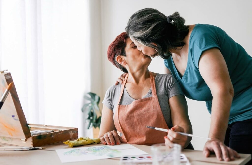 A couple exchanges a kiss while working on a painting together.