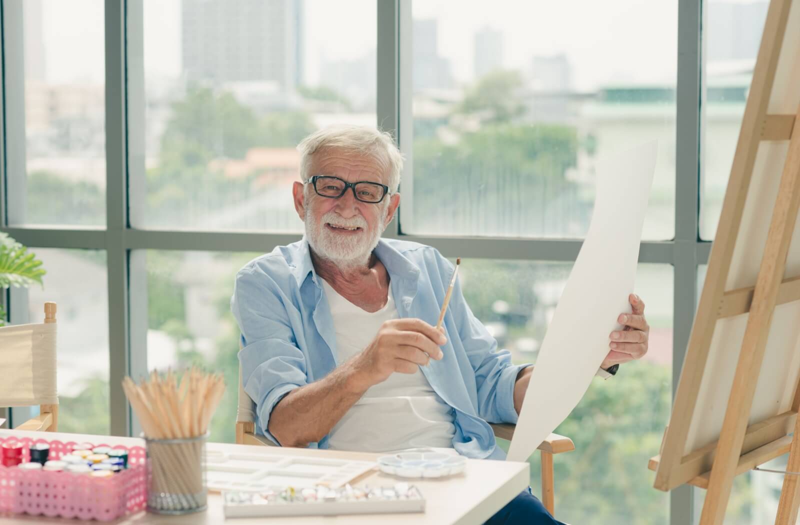Older adult painting in a bright studio, holding a brush and canvas with art supplies on the table.
