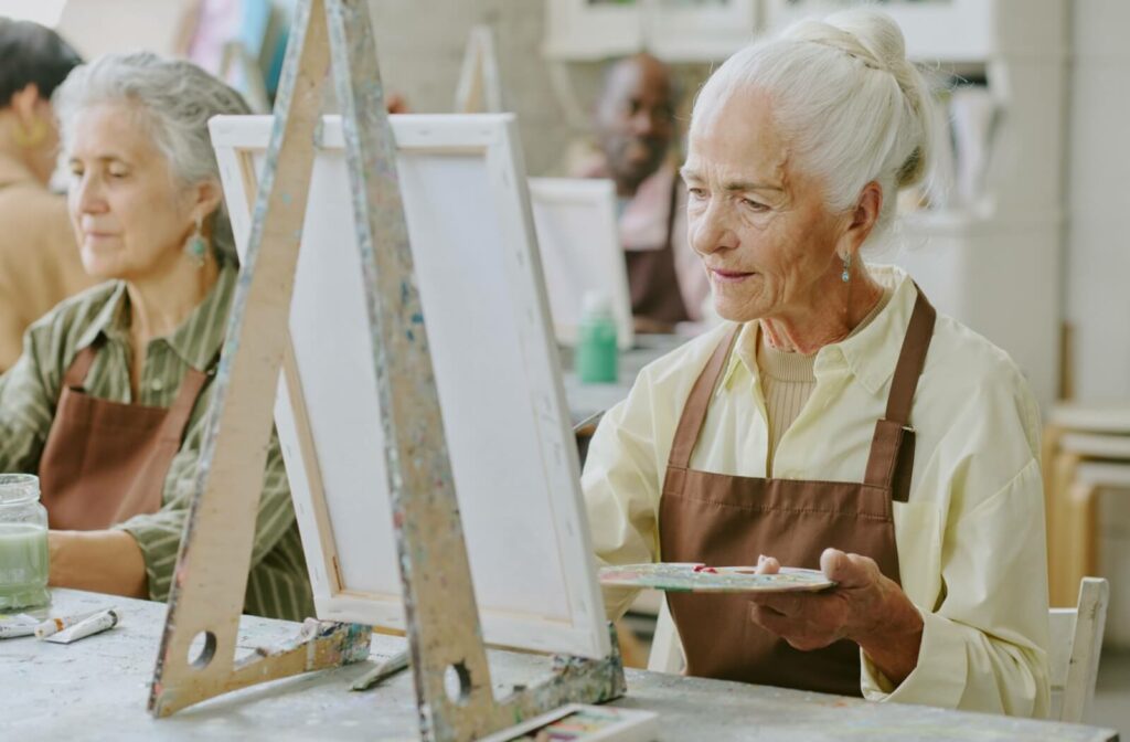Older adult painting in a group art class, holding a palette and brush while focusing on their work.