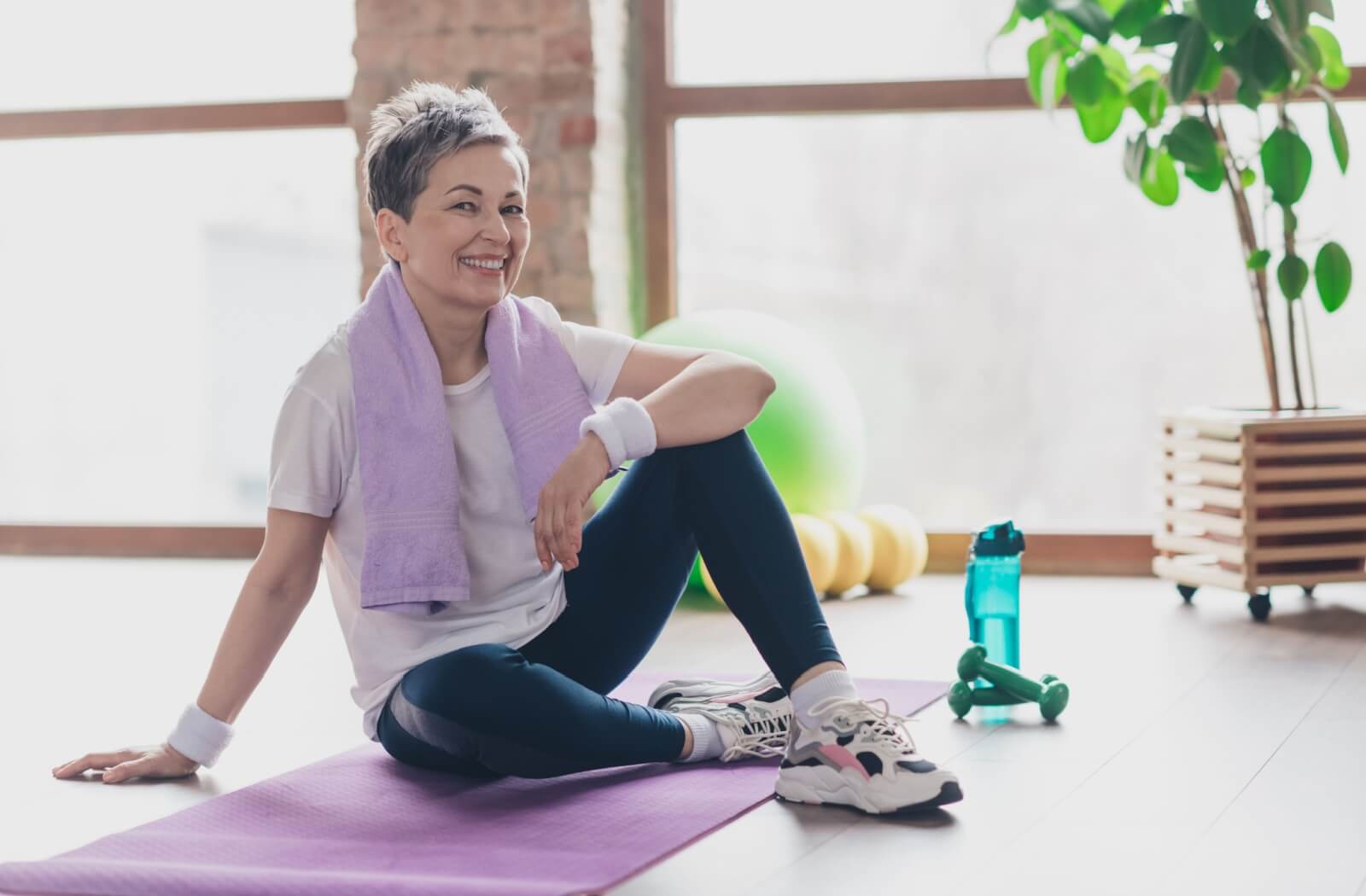 Smiling older adult sitting on a yoga mat with exercise equipment in a bright, indoor space.