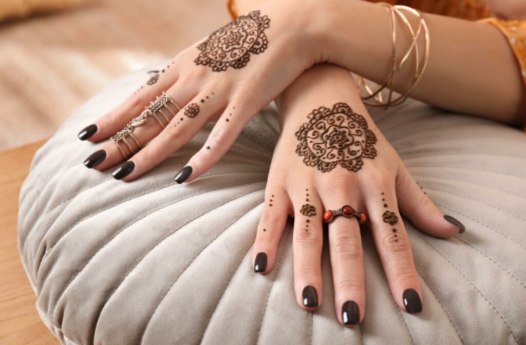 A person with elaborate, beautiful henna tattoos displays their hands on a grey suede cushion.