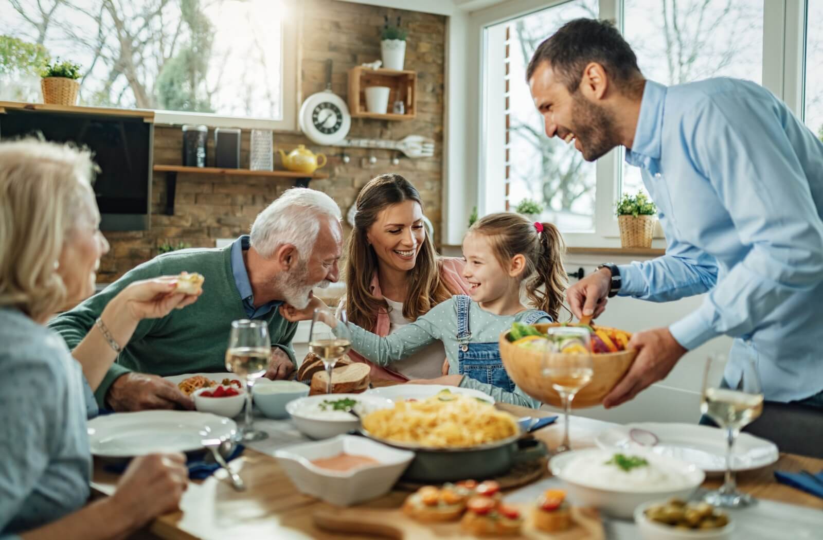 An older adult couple laughs with their adult children while playing with their grandchild at a table full of delicious food.
