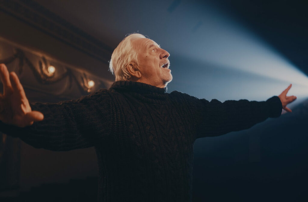 An older adult in a black sweater flourishes their arms on stage and recites their lines as a spotlight highlights their face.